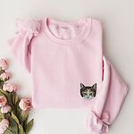 Light Pink crewneck sweatshirt laid flat beside pink peonies, with a calico cat face with green eyes embroidered on the left chest