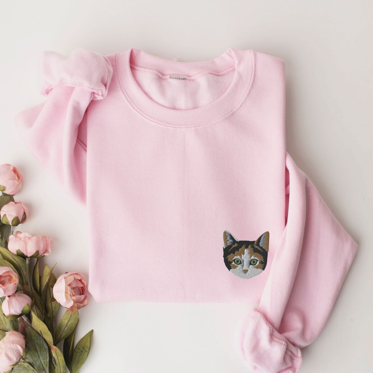 Calico Cat Sweatshirt Light Pink Light Pink crewneck sweatshirt laid flat beside pink peonies, with a calico cat face with green eyes embroidered on the left chest