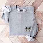 Sport Grey crewneck sweatshirt folded on a herringbone wood floor, with a calico cat face with green eyes embroidered on the left chest
