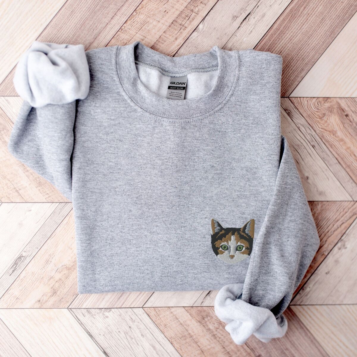 Calico Cat Sweatshirt Sport Grey Sport Grey crewneck sweatshirt folded on a herringbone wood floor, with a calico cat face with green eyes embroidered on the left chest