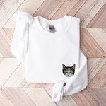 White crewneck sweatshirt folded on a herringbone wood floor, with a calico cat face with green eyes embroidered on the left chest