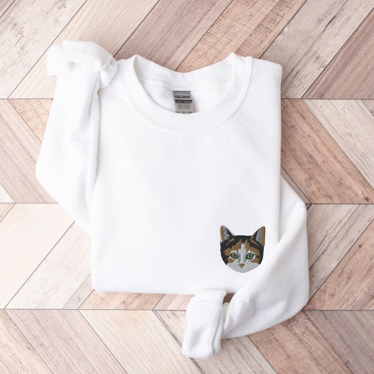 Calico Cat Sweatshirt White White crewneck sweatshirt folded on a herringbone wood floor, with a calico cat face with green eyes embroidered on the left chest
