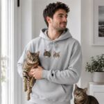 Man in a grey cat portrait hoodie holding his tabby cat, with a second tabby cat at his feet