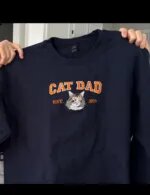 Navy cat dad sweatshirt with orange CAT DAD letters, an embroidered tabby and white cat face and EST. 2019
