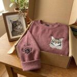 Embroidered cat memorial sweatshirt in a gift box beside a framed photo of the tabby cat with a halo
