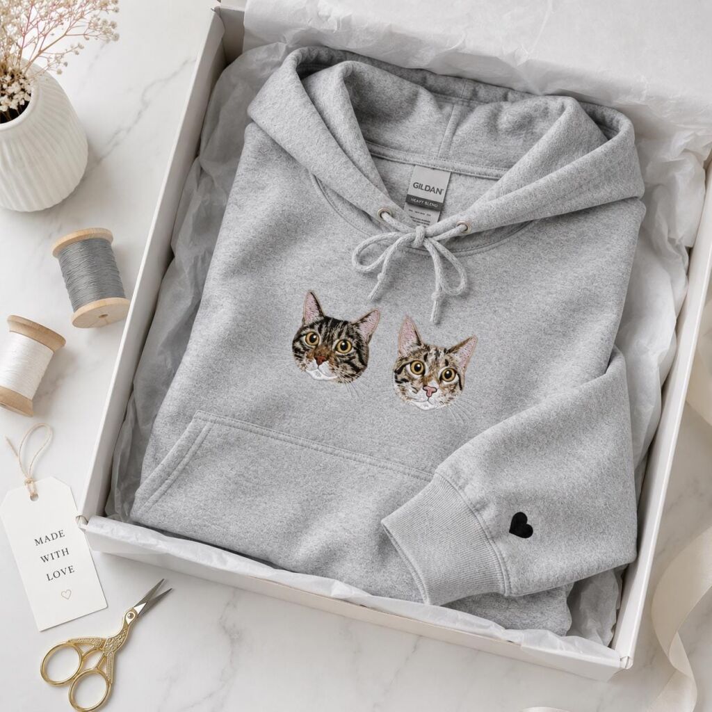 Cat memorial hoodie in light heather grey with two embroidered tabby cat portraits and a black heart on the sleeve, folded in a gift box