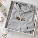 Cat memorial hoodie in light heather grey with two embroidered tabby cat portraits and a black heart on the sleeve, folded in a gift box