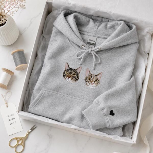 Cat memorial hoodie in light heather grey with two embroidered tabby cat portraits and a black heart on the sleeve, folded in a gift box