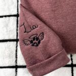 Sleeve embroidery of the name Lia in black script above an angel-wing paw print with a halo