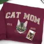 Maroon sweatshirt with CAT MOM varsity letters, an embroidered green-eyed tabby cat portrait and EST. 2023, next to the original cat photo