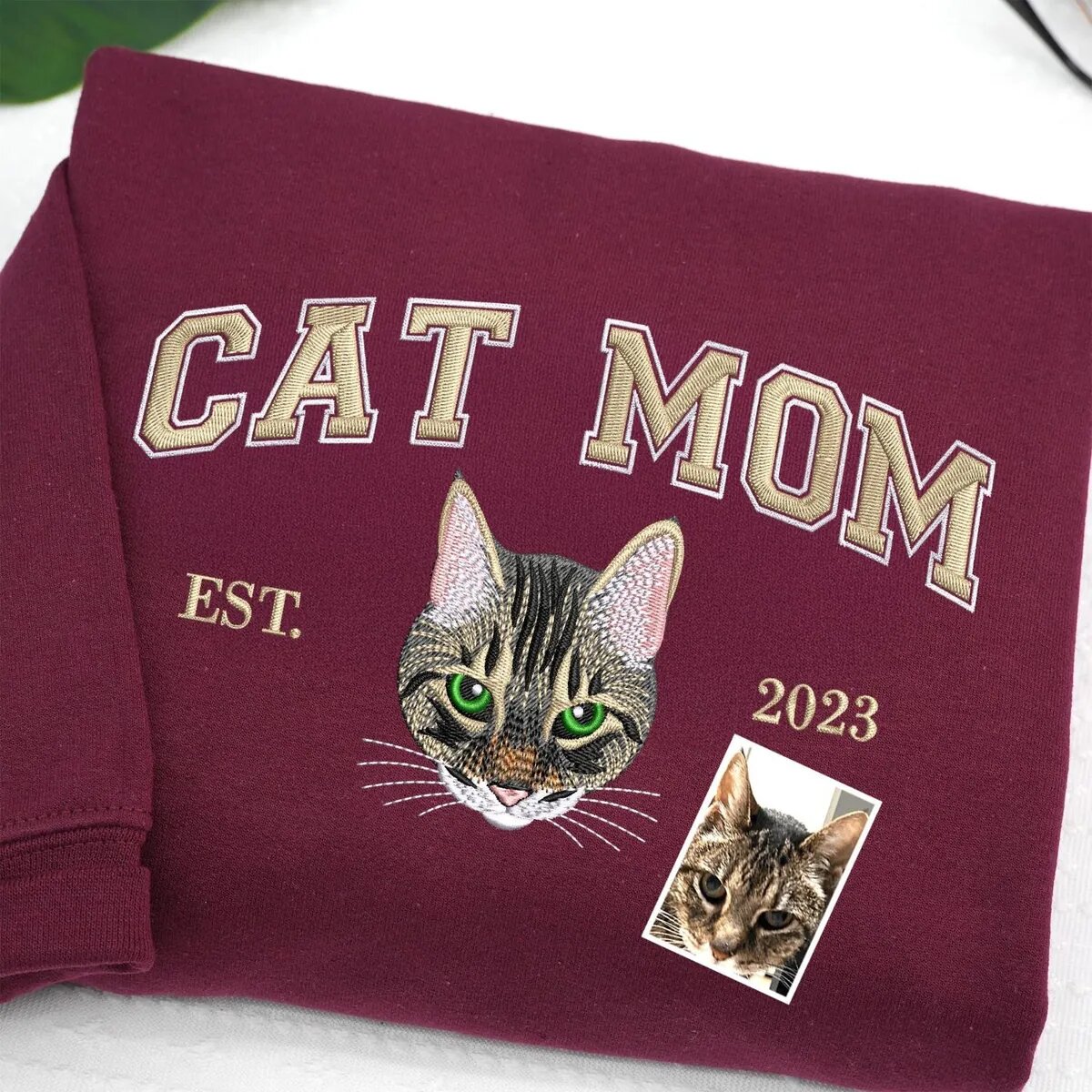 Cat Mom Varsity Sweatshirt – Maroon Maroon sweatshirt with CAT MOM varsity letters, an embroidered green-eyed tabby cat portrait and EST. 2023, next to the original cat photo