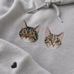 Close-up of two embroidered tabby cat faces showing the stitched fur, amber eyes and white whiskers