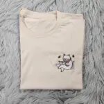 Cat reading book sweatshirt design shown on a cream T-shirt, a small embroidered white cat in a pink scarf reading beside a coffee cup on the left chest