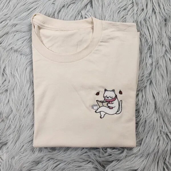 Cat reading book sweatshirt design shown on a cream T-shirt, a small embroidered white cat in a pink scarf reading beside a coffee cup on the left chest