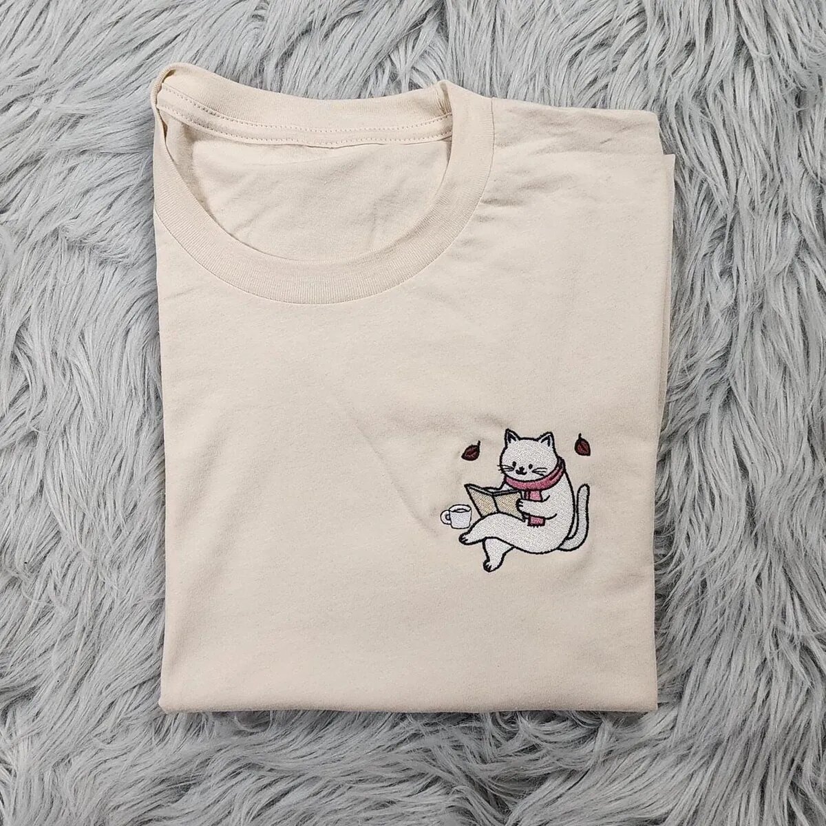 Cat Reading Book Shirt – Cream Cat reading book sweatshirt design shown on a cream T-shirt, a small embroidered white cat in a pink scarf reading beside a coffee cup on the left chest