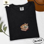 Black Comfort Colors T-shirt embroidered with an orange tabby cat asleep on a stack of green, brown and rust red books