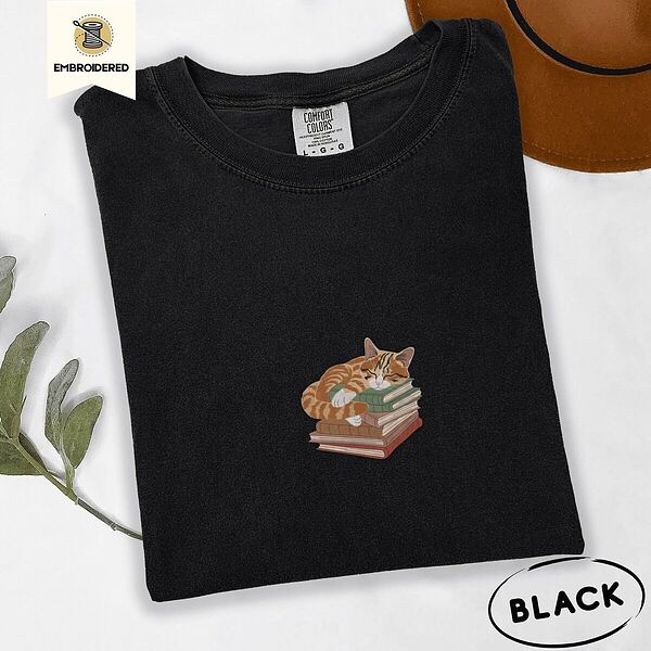 Black Comfort Colors T-shirt embroidered with an orange tabby cat asleep on a stack of green, brown and rust red books