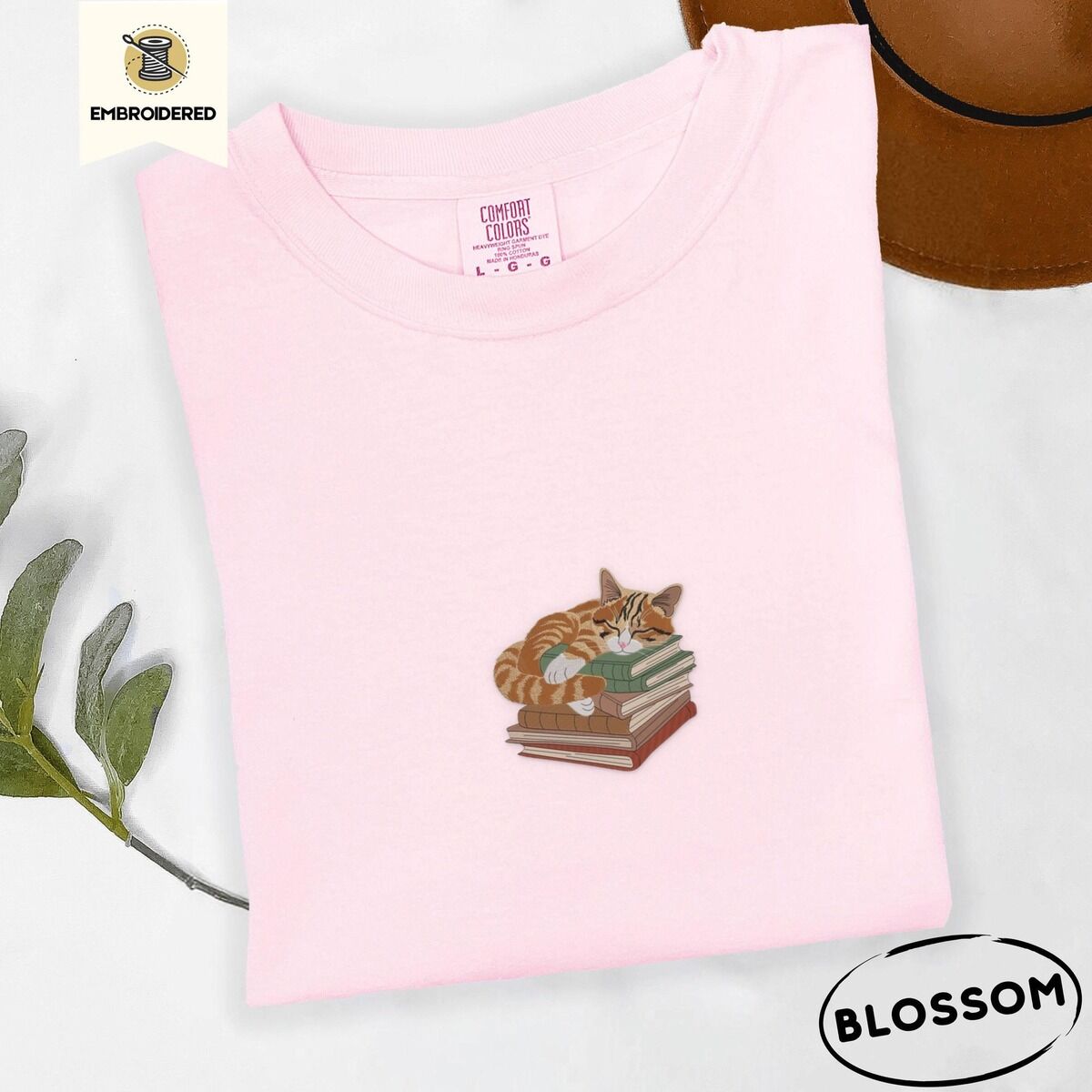 Cat Sleeping on Books Shirt – Blossom Blossom Comfort Colors T-shirt embroidered with an orange tabby cat asleep on a stack of green, brown and rust red books