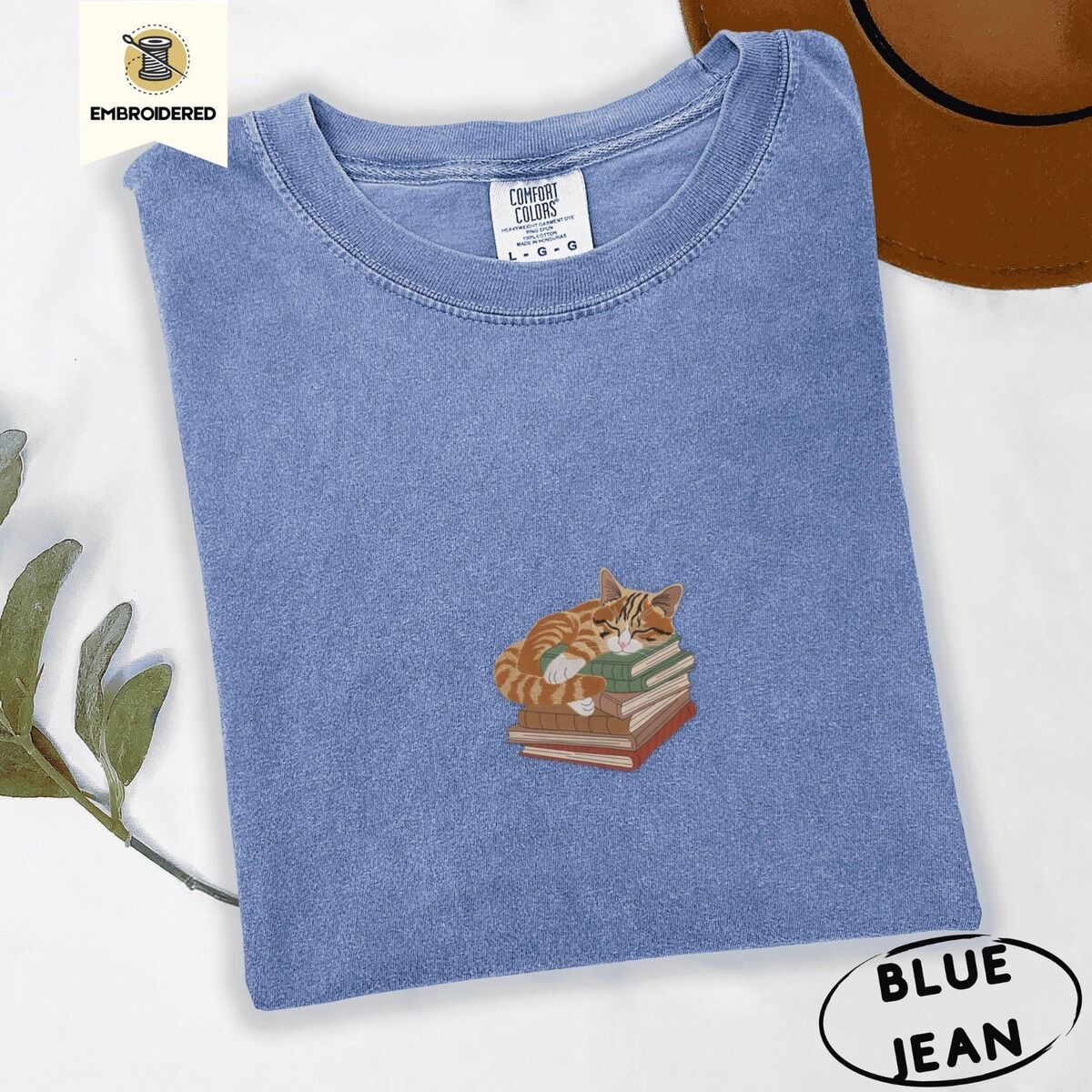 Cat Sleeping on Books Shirt – Blue Jean Blue Jean Comfort Colors T-shirt embroidered with an orange tabby cat asleep on a stack of green, brown and rust red books
