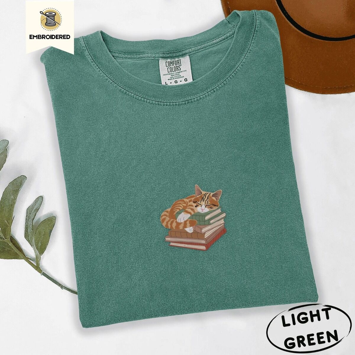 Cat Sleeping on Books Shirt – Light Green Light Green Comfort Colors T-shirt embroidered with an orange tabby cat asleep on a stack of green, brown and rust red books