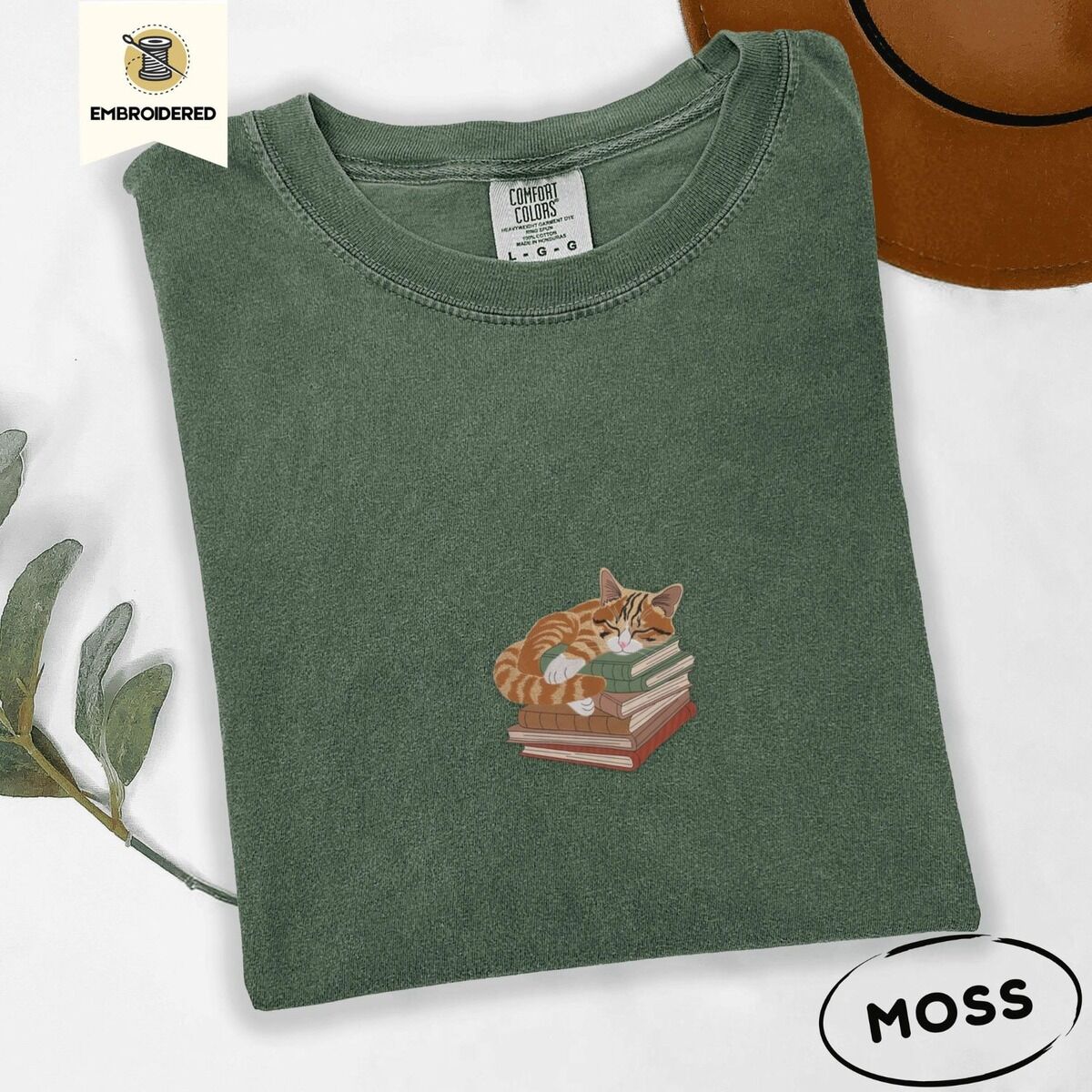 Cat Sleeping on Books Shirt – Moss Moss Comfort Colors T-shirt embroidered with an orange tabby cat asleep on a stack of green, brown and rust red books