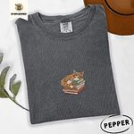 Pepper Comfort Colors T-shirt embroidered with an orange tabby cat asleep on a stack of green, brown and rust red books