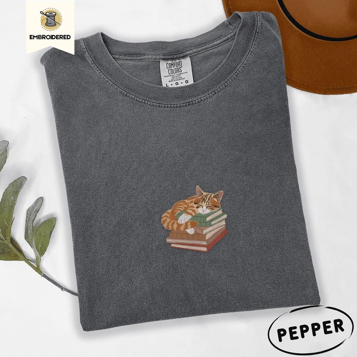 Cat Sleeping on Books Shirt – Pepper Pepper Comfort Colors T-shirt embroidered with an orange tabby cat asleep on a stack of green, brown and rust red books
