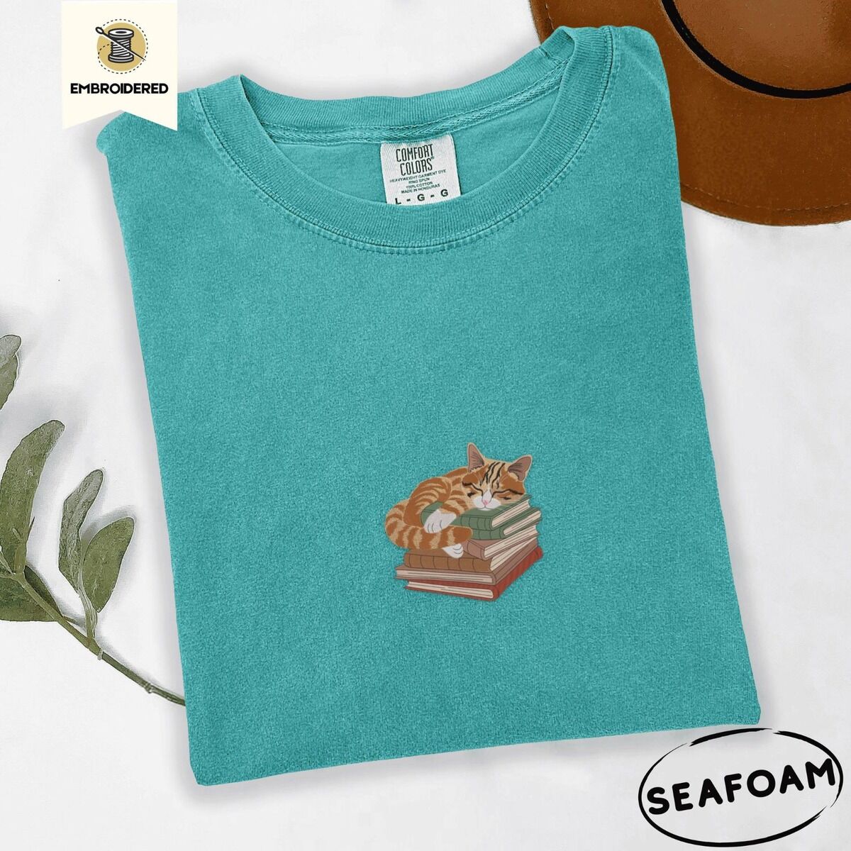 Cat Sleeping on Books Shirt – Seafoam Seafoam Comfort Colors T-shirt embroidered with an orange tabby cat asleep on a stack of green, brown and rust red books