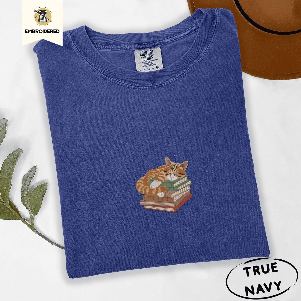 Cat Sleeping on Books Shirt – True Navy True Navy Comfort Colors T-shirt embroidered with an orange tabby cat asleep on a stack of green, brown and rust red books