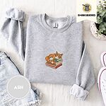 Ash crewneck sweatshirt folded with denim shorts, white sneakers and a potted plant, embroidered with an orange tabby cat asleep on a stack of green, brown and red books