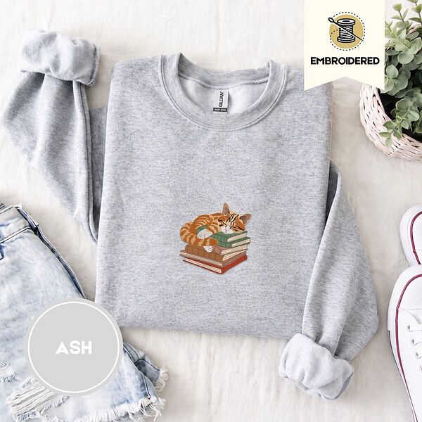 Ash crewneck sweatshirt folded with denim shorts, white sneakers and a potted plant, embroidered with an orange tabby cat asleep on a stack of green, brown and red books