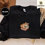Black crewneck sweatshirt with a brown felt hat and dried bunny tail grass, embroidered with an orange tabby cat asleep on a stack of green, brown and red books