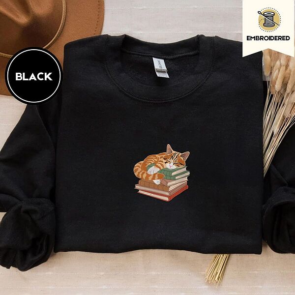 Black crewneck sweatshirt with a brown felt hat and dried bunny tail grass, embroidered with an orange tabby cat asleep on a stack of green, brown and red books