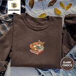 Dark Chocolate crewneck sweatshirt on a plaid flannel shirt with yellow autumn leaves and denim shorts, embroidered with an orange tabby cat asleep on a stack of green, brown and red books