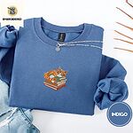 Indigo Blue crewneck sweatshirt folded with light jeans and dried twigs, embroidered with an orange tabby cat asleep on a stack of green, brown and red books