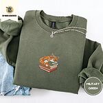Military Green crewneck sweatshirt folded with light jeans and a silver chain necklace, embroidered with an orange tabby cat asleep on a stack of green, brown and red books