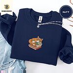 Navy crewneck sweatshirt folded with ripped jeans and a silver chain necklace, embroidered with an orange tabby cat asleep on a stack of green, brown and red books