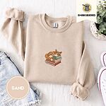 Sand crewneck sweatshirt folded with denim shorts, white sneakers and a potted plant, embroidered with an orange tabby cat asleep on a stack of green, brown and red books