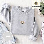 Ash crewneck sweatshirt folded with denim shorts, white sneakers and a potted plant, embroidered with a mini Cavachon face in cream and apricot with a white muzzle