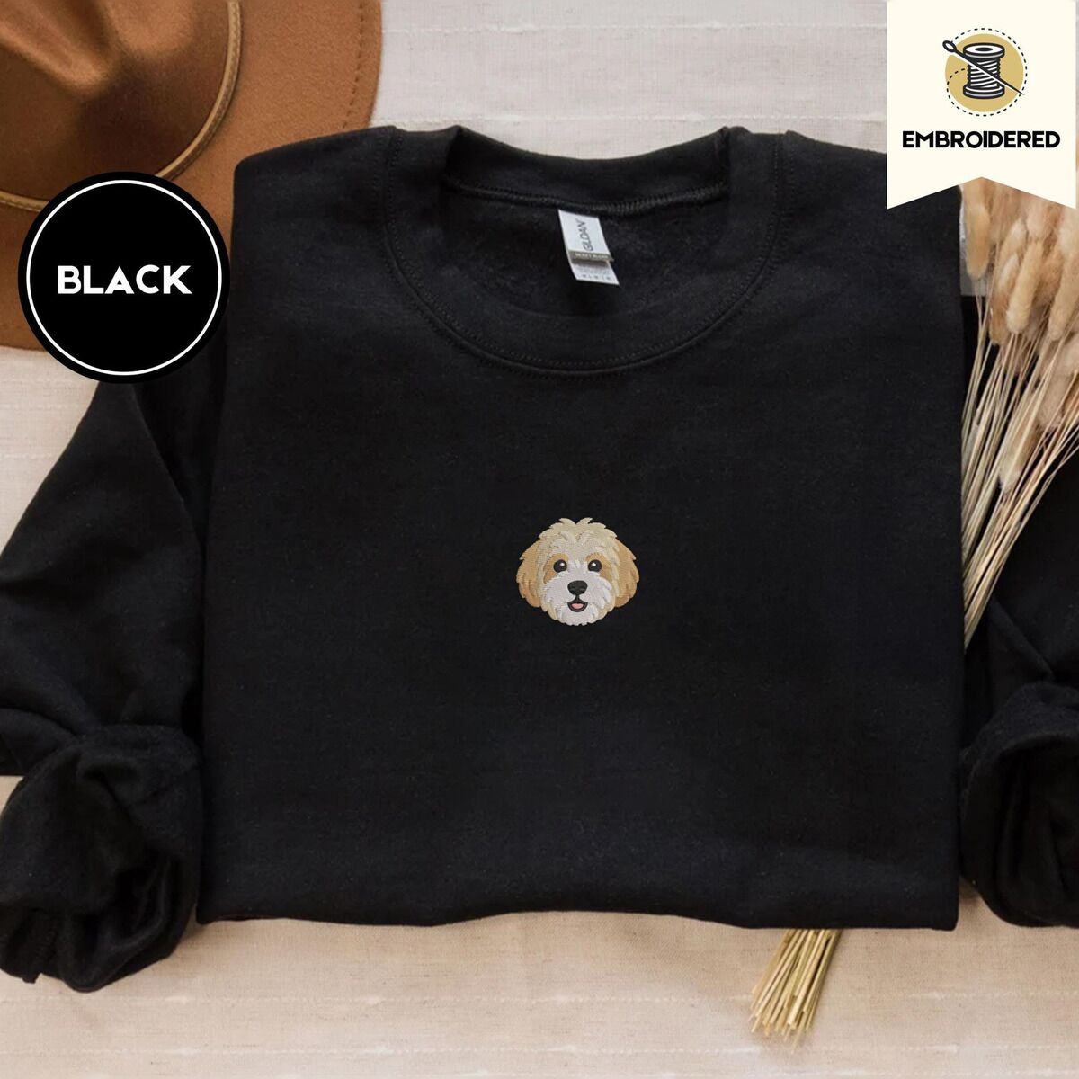 Cavachon Sweatshirt – Black Black crewneck sweatshirt with a brown felt hat and dried bunny tail grass, embroidered with a mini Cavachon face in cream and apricot with a white muzzle