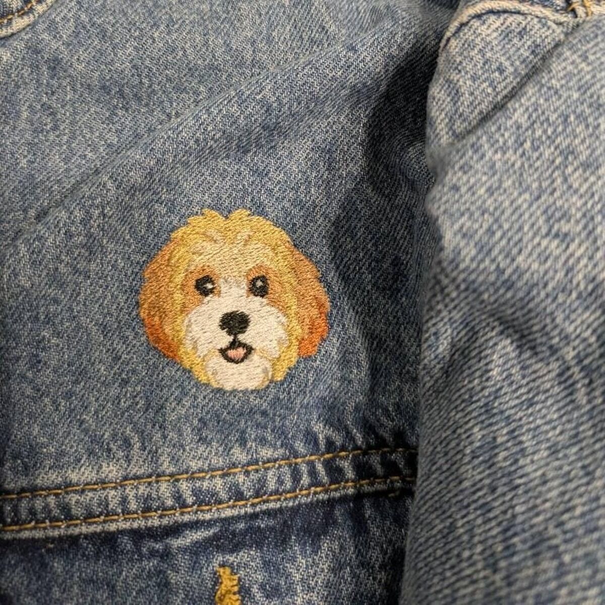 Cavachon Face Embroidery Close-Up Close-up of the real stitched mini Cavachon face in cream and apricot with a white muzzle, black eyes and a pink tongue, shown on a denim jacket