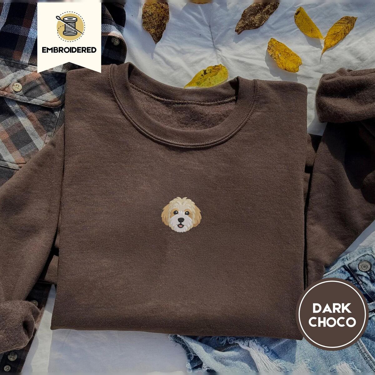 Cavachon Sweatshirt – Dark Chocolate Dark Chocolate crewneck sweatshirt on a plaid flannel shirt with yellow autumn leaves and denim shorts, embroidered with a mini Cavachon face in cream and apricot with a white muzzle