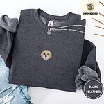 Dark Heather crewneck sweatshirt folded with ripped jeans and a silver chain necklace, embroidered with a mini Cavachon face in cream and apricot with a white muzzle