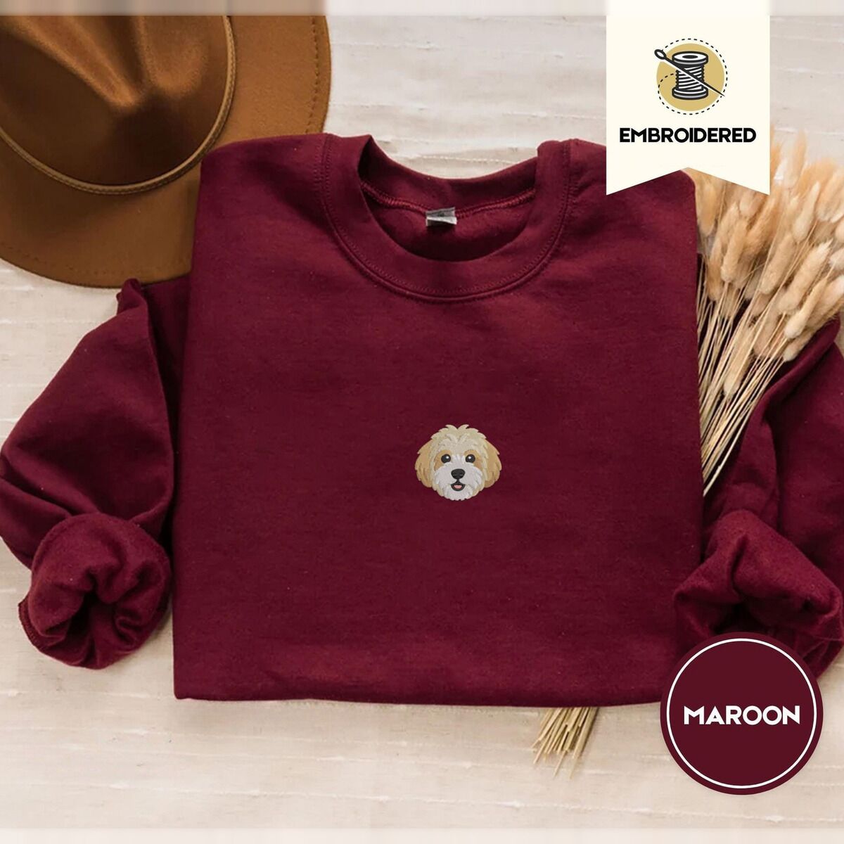 Cavachon Sweatshirt – Maroon Maroon crewneck sweatshirt with a brown felt hat and dried bunny tail grass, embroidered with a mini Cavachon face in cream and apricot with a white muzzle