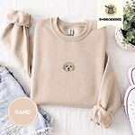 Sand crewneck sweatshirt folded with denim shorts, white sneakers and a potted plant, embroidered with a mini Cavachon face in cream and apricot with a white muzzle