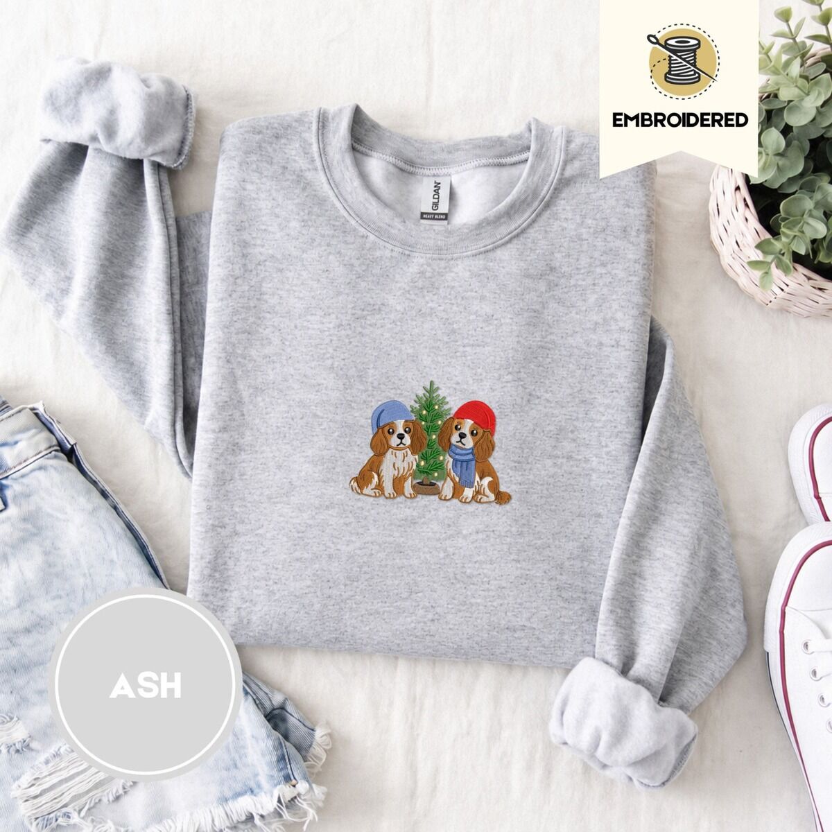 Cavalier King Charles Spaniel Christmas Sweatshirt – Ash Ash crewneck sweatshirt folded with denim shorts, white sneakers and a potted plant, embroidered with two Cavalier King Charles Spaniels in knit beanies beside a little lit Christmas tree