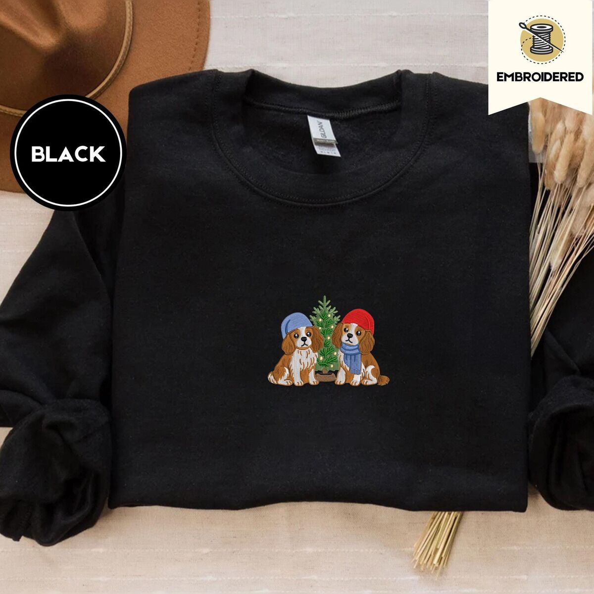 Cavalier King Charles Spaniel Christmas Sweatshirt – Black Black crewneck sweatshirt with a brown felt hat and dried bunny tail grass, embroidered with two Cavalier King Charles Spaniels in knit beanies beside a little lit Christmas tree