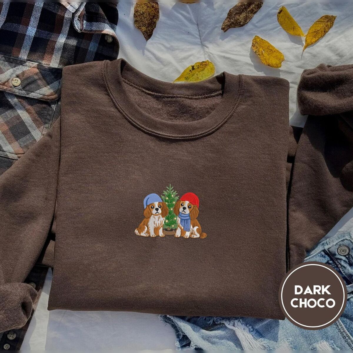 Cavalier King Charles Spaniel Christmas Sweatshirt – Dark Chocolate Dark Chocolate crewneck sweatshirt on a plaid flannel shirt with yellow autumn leaves and denim shorts, embroidered with two Cavalier King Charles Spaniels in knit beanies beside a little lit Christmas tree