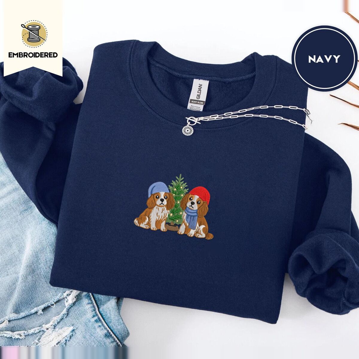 Cavalier King Charles Spaniel Christmas Sweatshirt – Navy Navy crewneck sweatshirt folded with ripped jeans and a silver chain necklace, embroidered with two Cavalier King Charles Spaniels in knit beanies beside a little lit Christmas tree