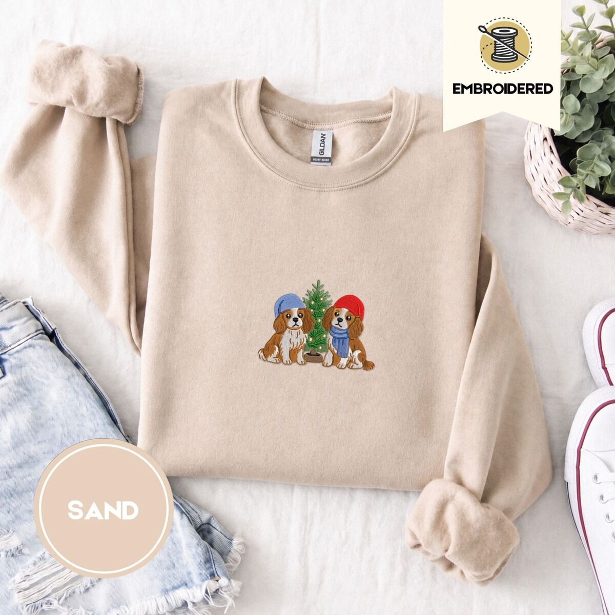 Cavalier King Charles Spaniel Christmas Sweatshirt – Sand Sand crewneck sweatshirt folded with denim shorts, white sneakers and a potted plant, embroidered with two Cavalier King Charles Spaniels in knit beanies beside a little lit Christmas tree